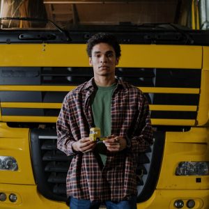 Adult male with afro hair stands holding a beverage in front of a large yellow truck.